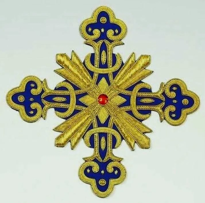 Gold and blue embroidered cross badge with red center gem on light background.